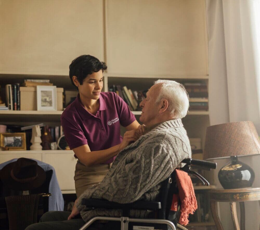A caregiver in a purple shirt assists an elderly man in a wheelchair inside a warmly lit room with bookshelves. - Home Instead