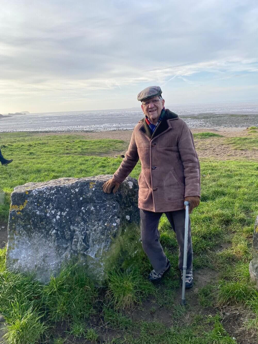 An elderly man wearing a coat and hat rests against a large rock with a cane, standing on a grassy field near the shore. - Home Instead