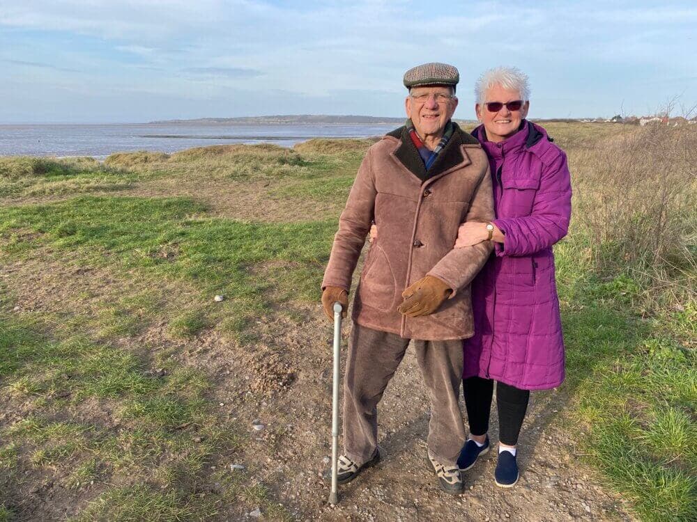 Elderly couple smiling and standing arm in arm on a grassy path near the sea on a clear day. - Home Instead