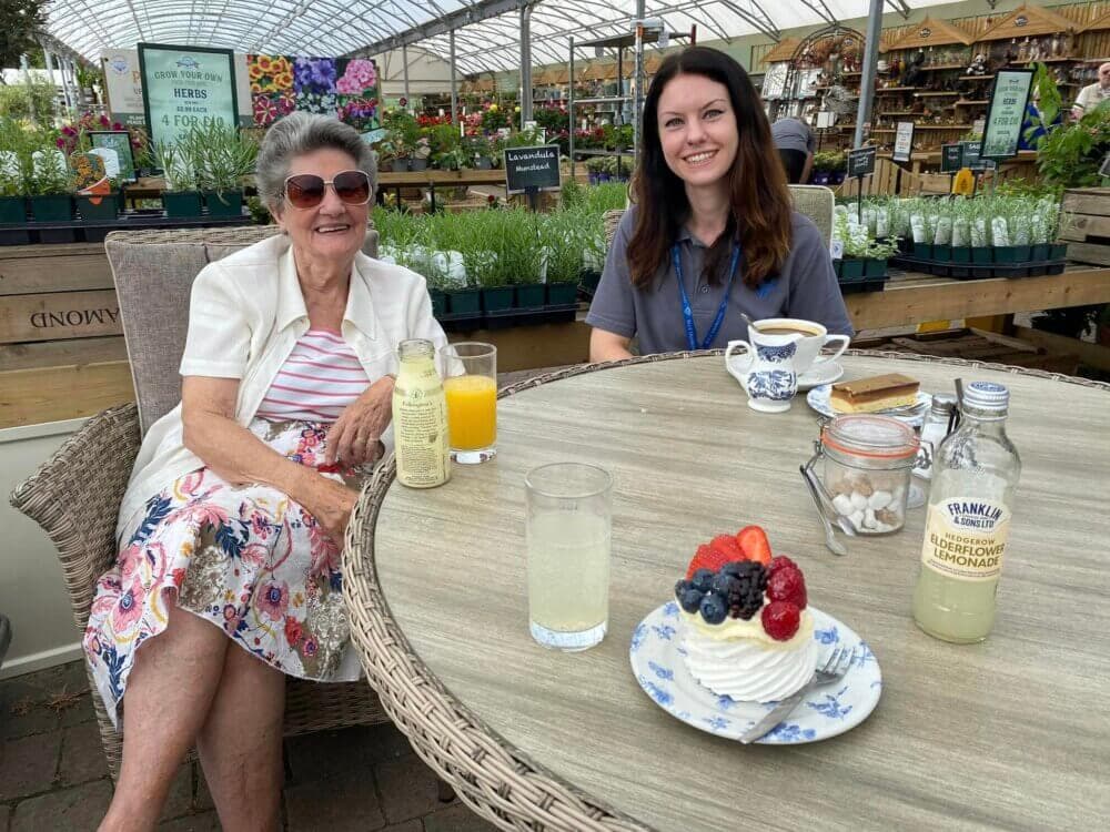 Two women sitting at a garden center café table with drinks and desserts, smiling. Plants and flowers in the background. - Home Instead