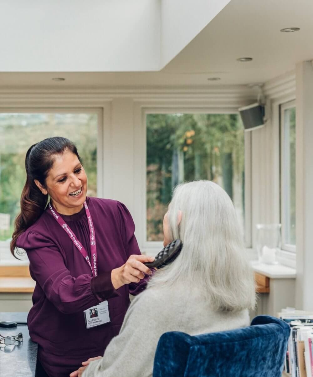 A caregiver in a burgundy shirt brushes the hair of an elderly woman seated in a blue chair in a sunny room. - Home Instead