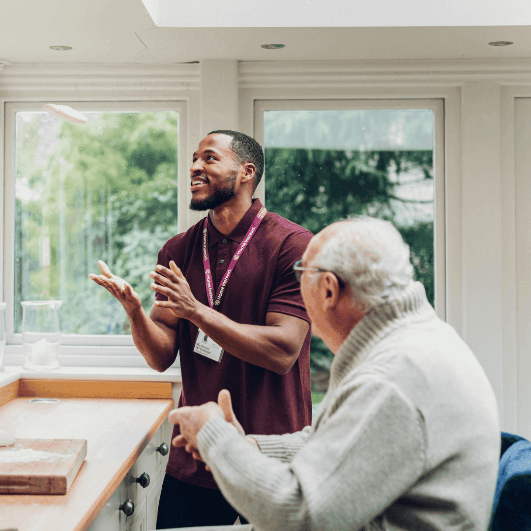 Younger man in a maroon polo shirt speaking to an older man in a sweater at a kitchen counter with large windows. - Home Instead