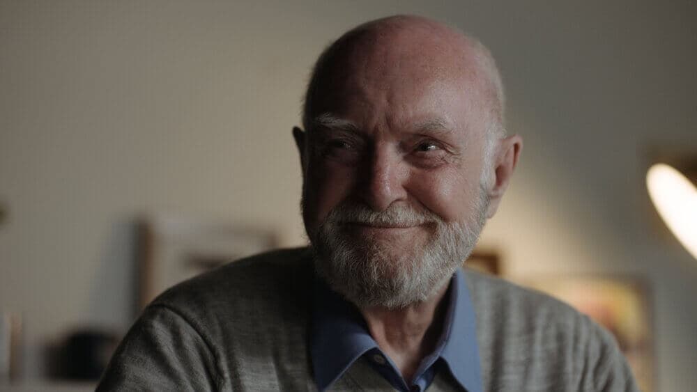 Elderly man with a bald head and a beard smiling warmly, wearing a collared shirt and a gray sweater in a dimly lit room. - Home Instead