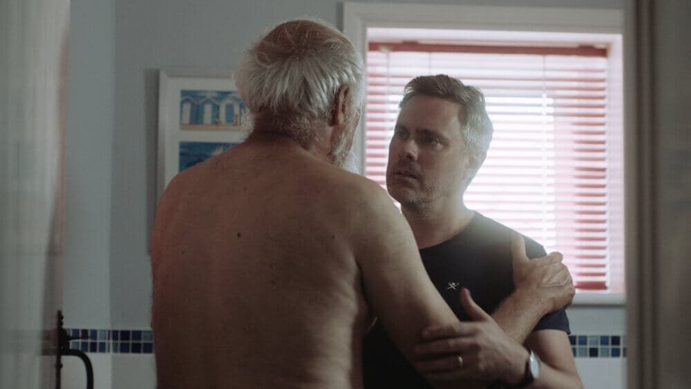 Two men, one shirtless and elderly with gray hair, the other younger in a black shirt, have a serious conversation. - Home Instead