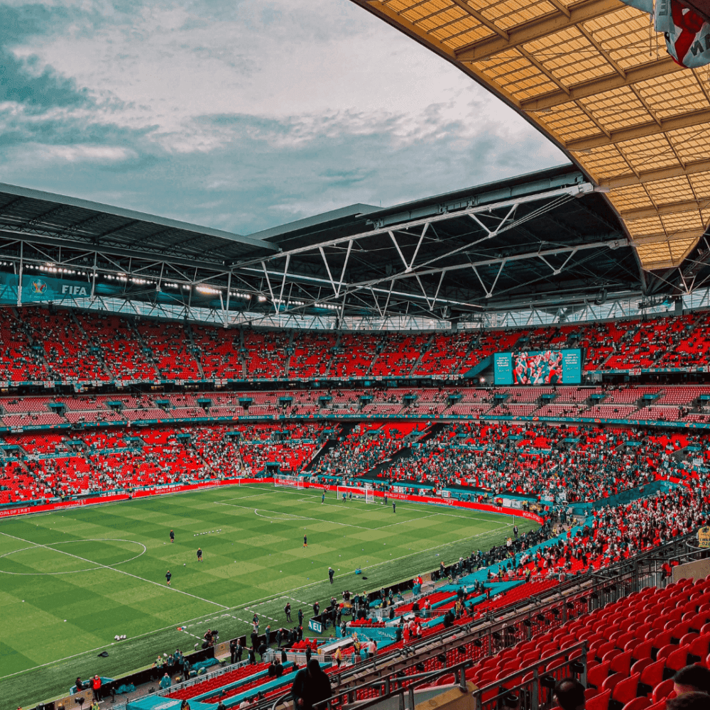 A large, open-roof stadium with a green soccer field, filled with spectators in red seats, under a cloudy sky. - Home Instead