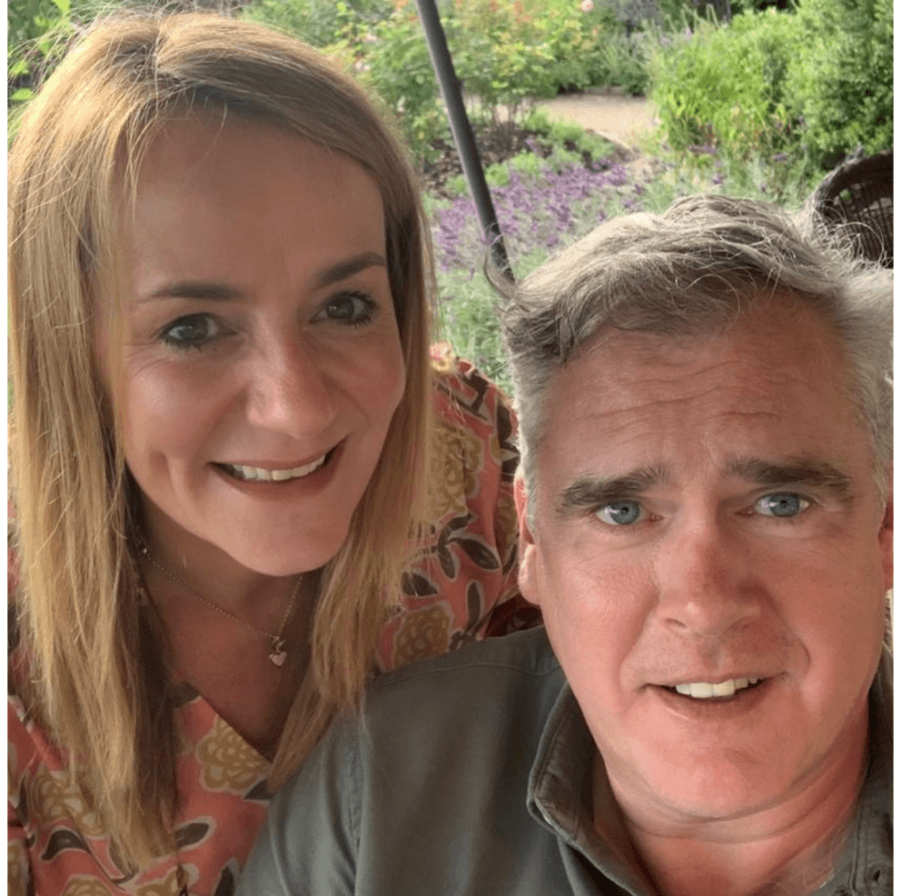 A woman with blonde hair and a man with gray hair smiling outdoors with greenery in the background. - Home Instead