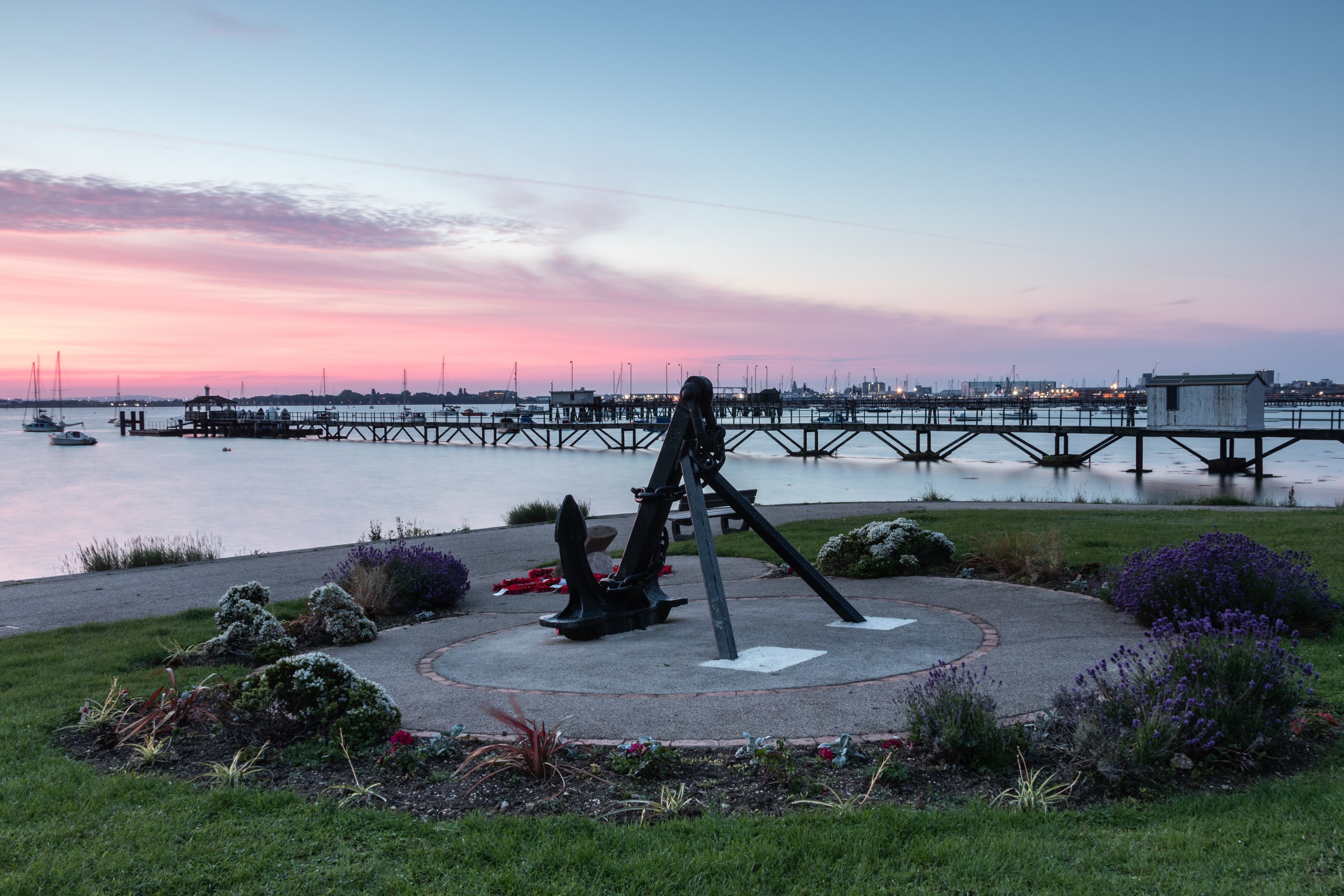 An anchor monument near a waterfront at sunset, with a pier and boats visible in the distance. - Home Instead