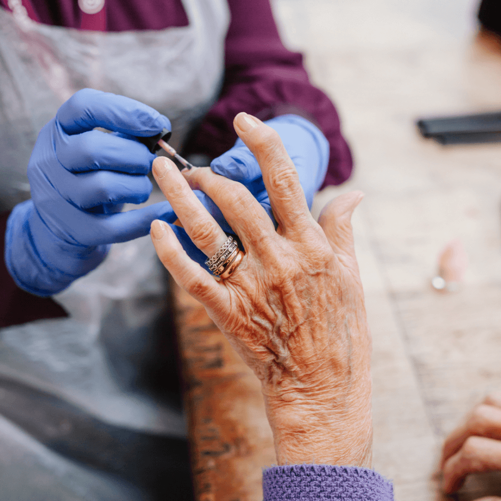 Close-up of an elderly person getting a manicure, with hands wearing blue gloves working on their nails. - Home Instead