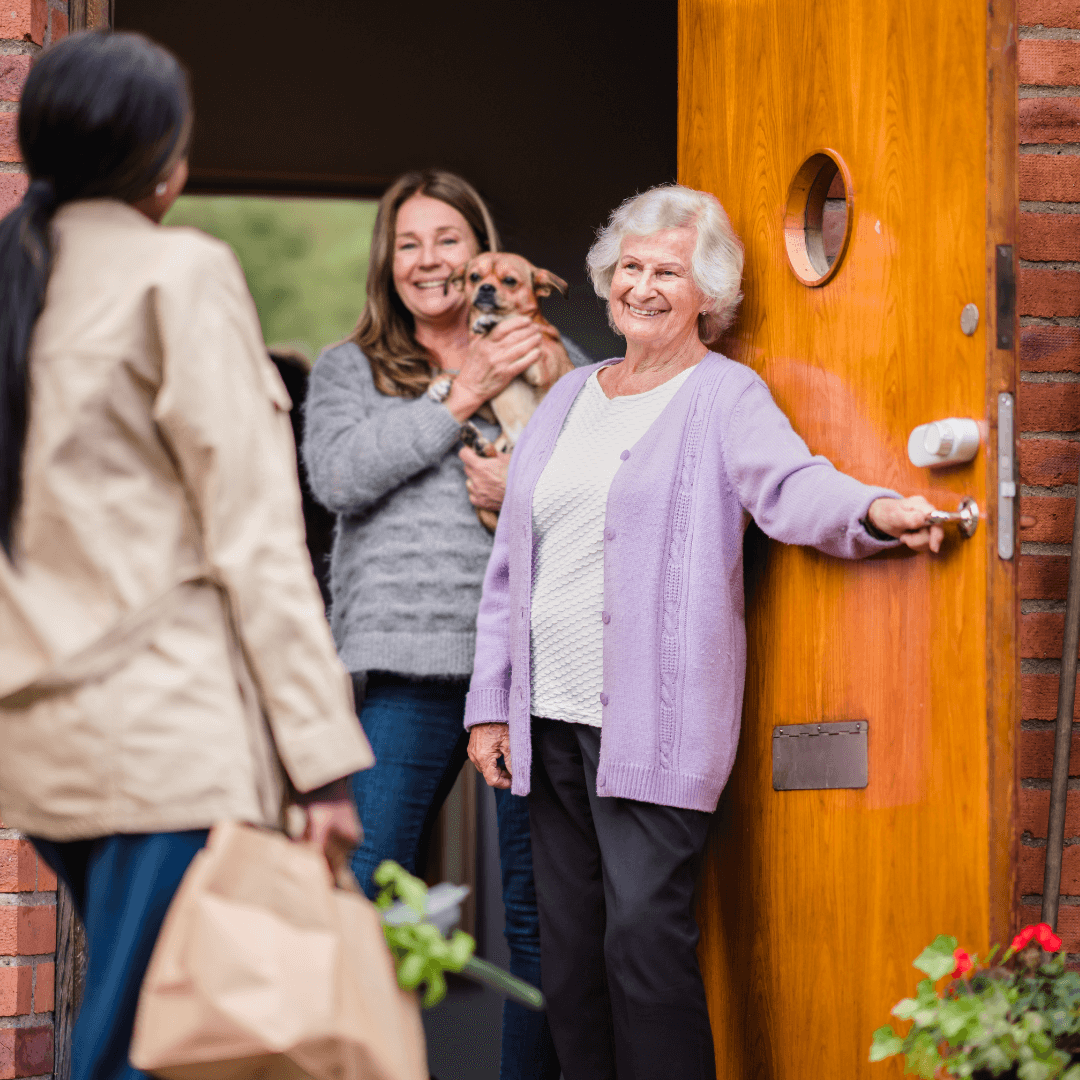 Woman at door with groceries greets smiling elderly woman and another woman holding a dog inside a home. - Home Instead