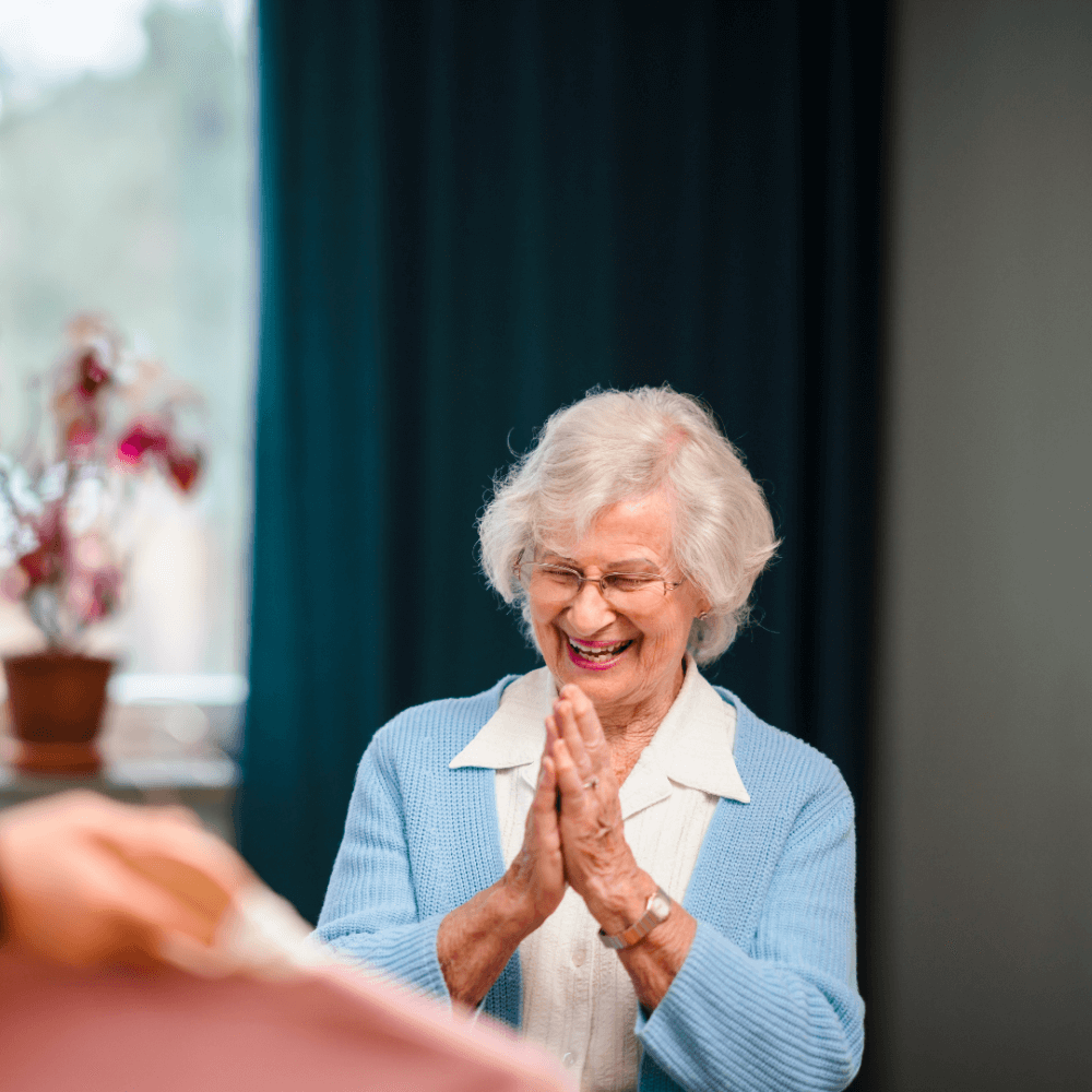 An elderly woman with white hair and a blue cardigan clasps her hands and smiles in a warmly lit room. - Home Instead