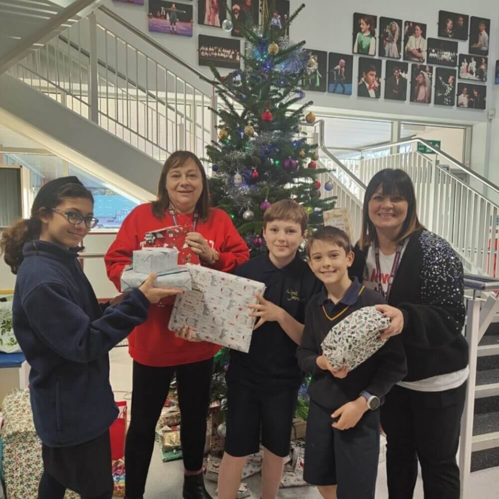 Four children and two adults holding presents in front of a decorated Christmas tree in a school setting. - Home Instead