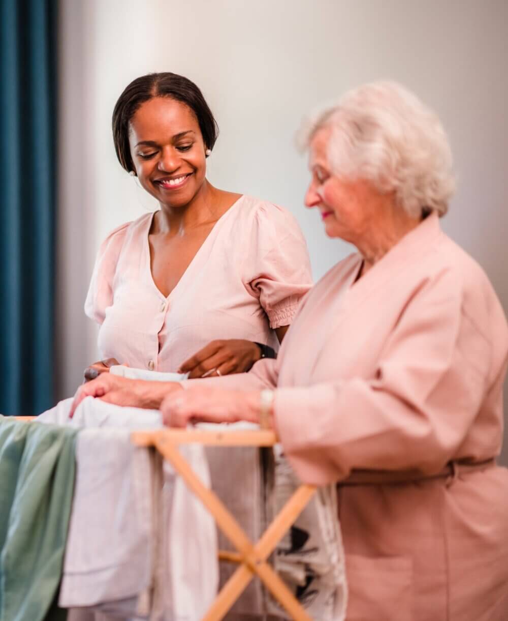 A young woman smiling while helping an elderly woman with laundry on a drying rack. - Home Instead