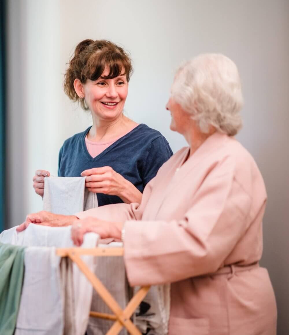 Home Instead Bournemouth & Christchurch Care Professional and client, smile and chat while hanging laundry together indoors.