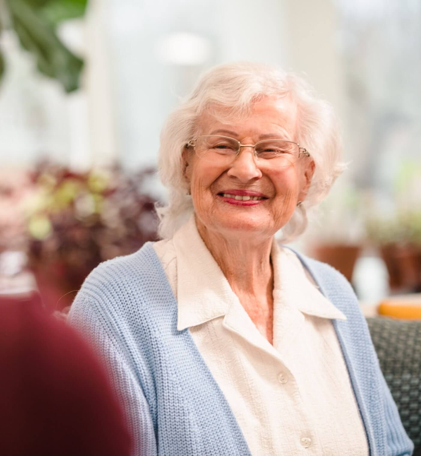 A smiling elderly client with white hair and glasses wears a light blue cardigan and white shirt, sitting indoors with a Home Instead Bournemouth & Christchurch Care Professional