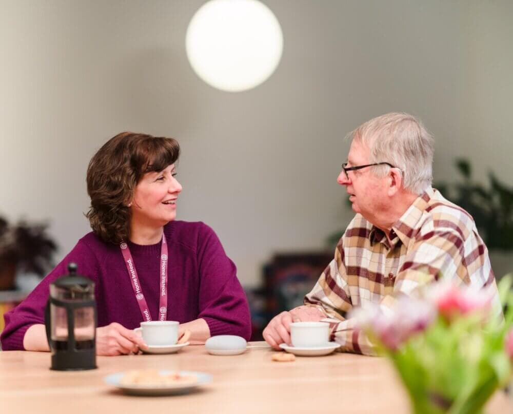 Two people sit at a table with coffee cups, smiling and talking. A French press is on the table. - Home Instead