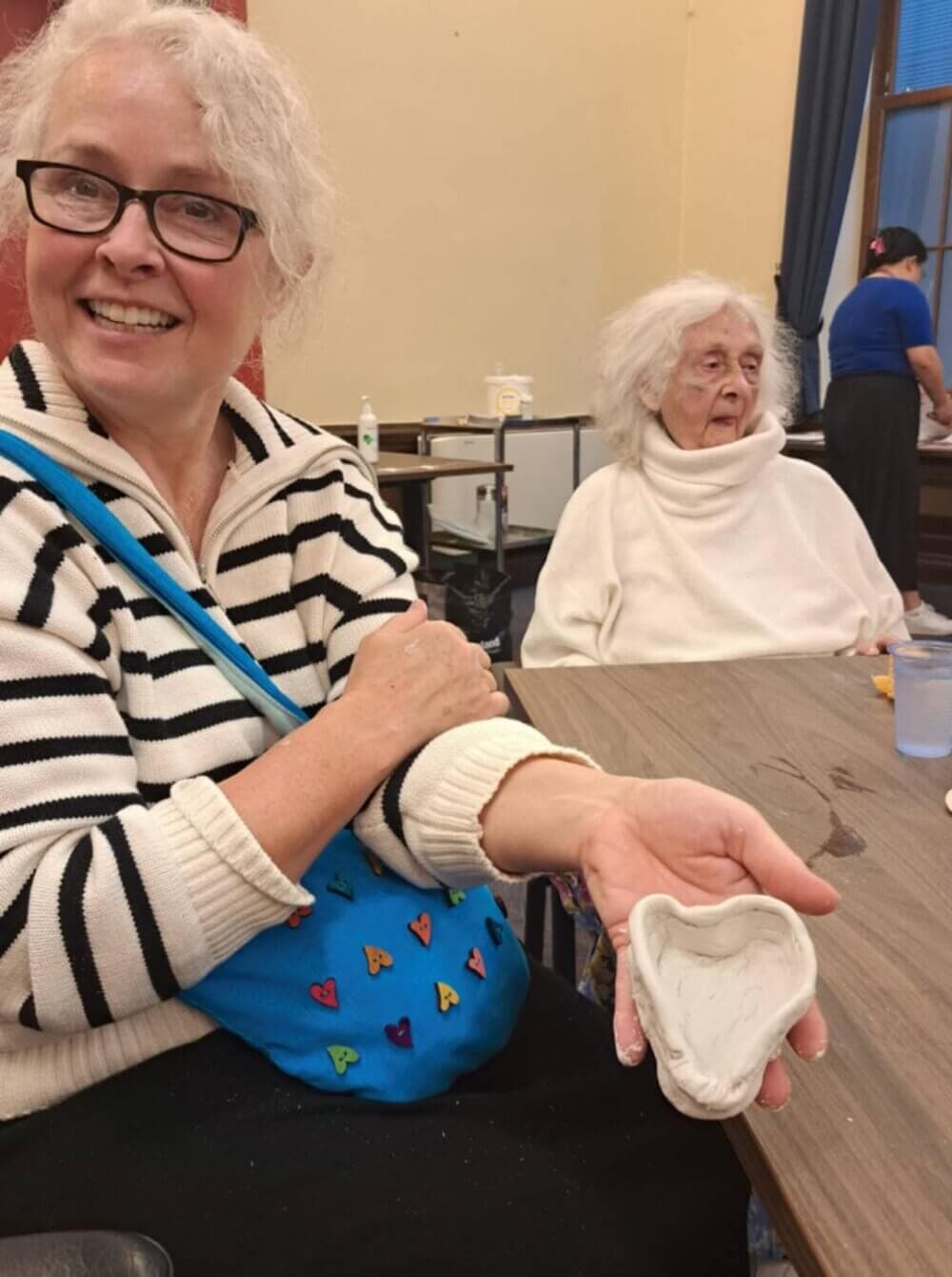 Two elderly women at a table; one smiles and holds up a heart-shaped clay craft. - Home Instead