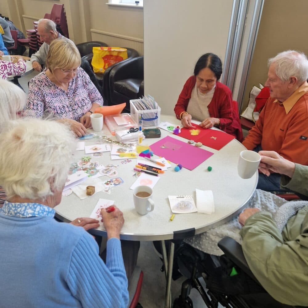 Five older adults sit around a table doing crafts together, with mugs and art supplies scattered on the table. - Home Instead