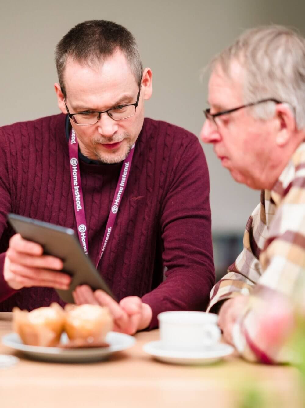 Two men sitting at a table, one showing the other something on a tablet, with muffins and a cup of coffee in front of them. - Home Instead