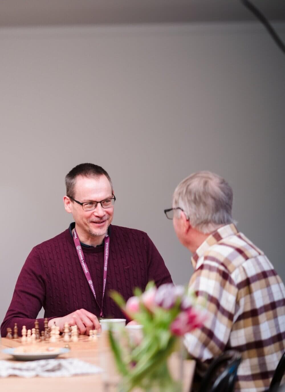 Two men are seated at a table with a chessboard, engaged in conversation. A vase with flowers is also on the table. - Home Instead