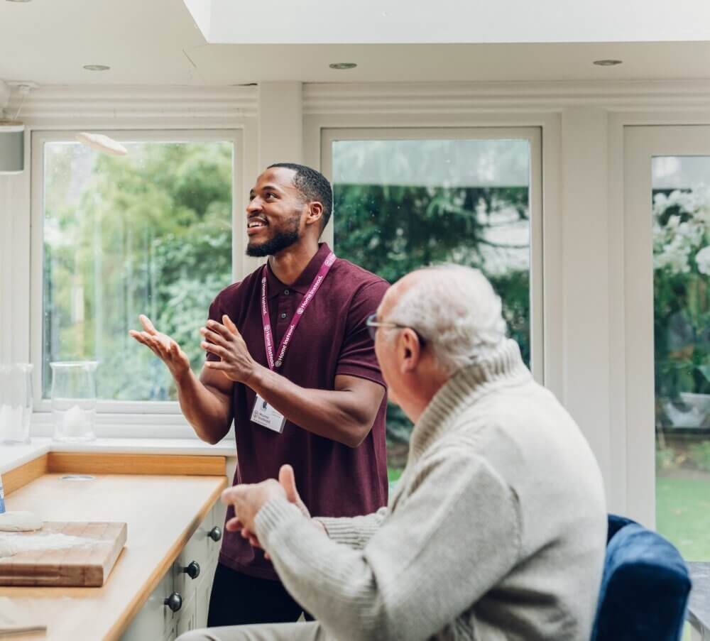 A man and an elderly person engage in conversation by a bright window. The man is smiling and gesturing with his hands. - Home Instead
