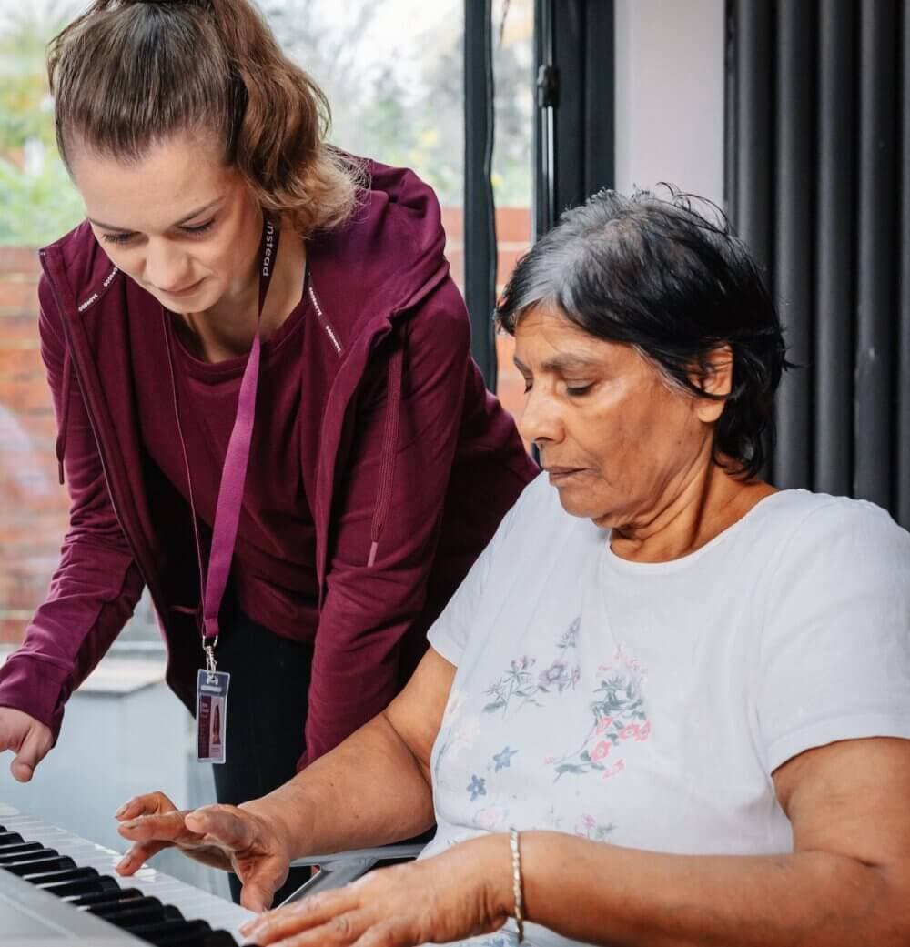 A young woman helps an elderly woman play the piano indoors. - Home Instead