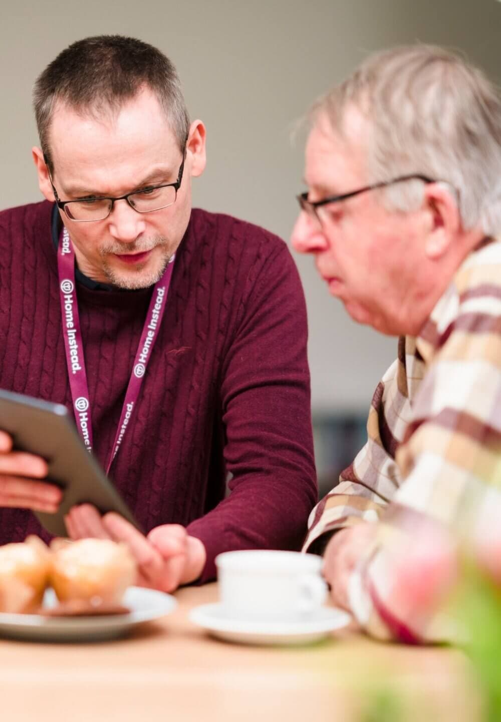 Two men, one with a name badge, look at a tablet at a table with coffee and pastries. - Home Instead