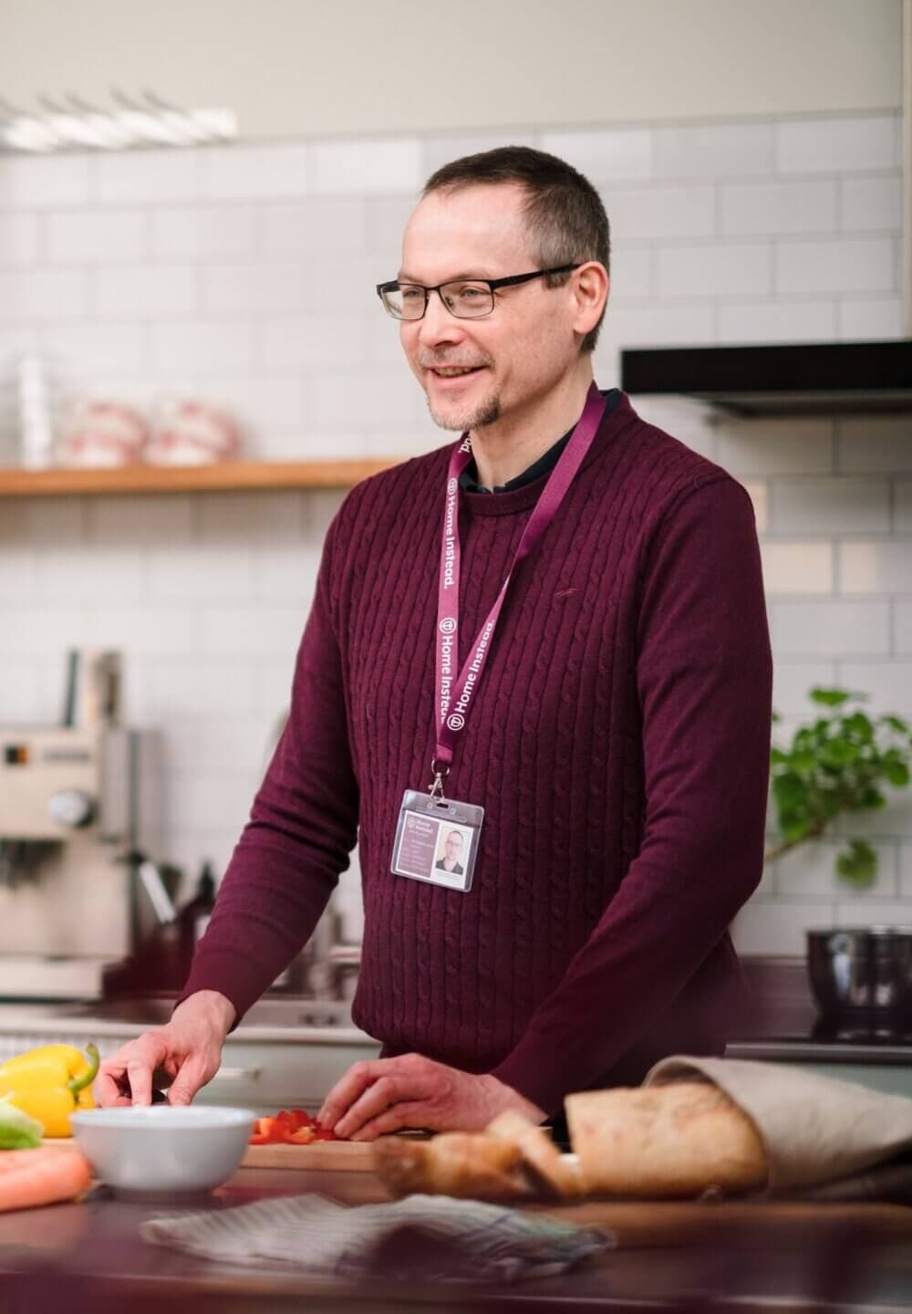 A man wearing glasses and a maroon sweater slices vegetables in a kitchen, with a nametag lanyard around his neck. - Home Instead