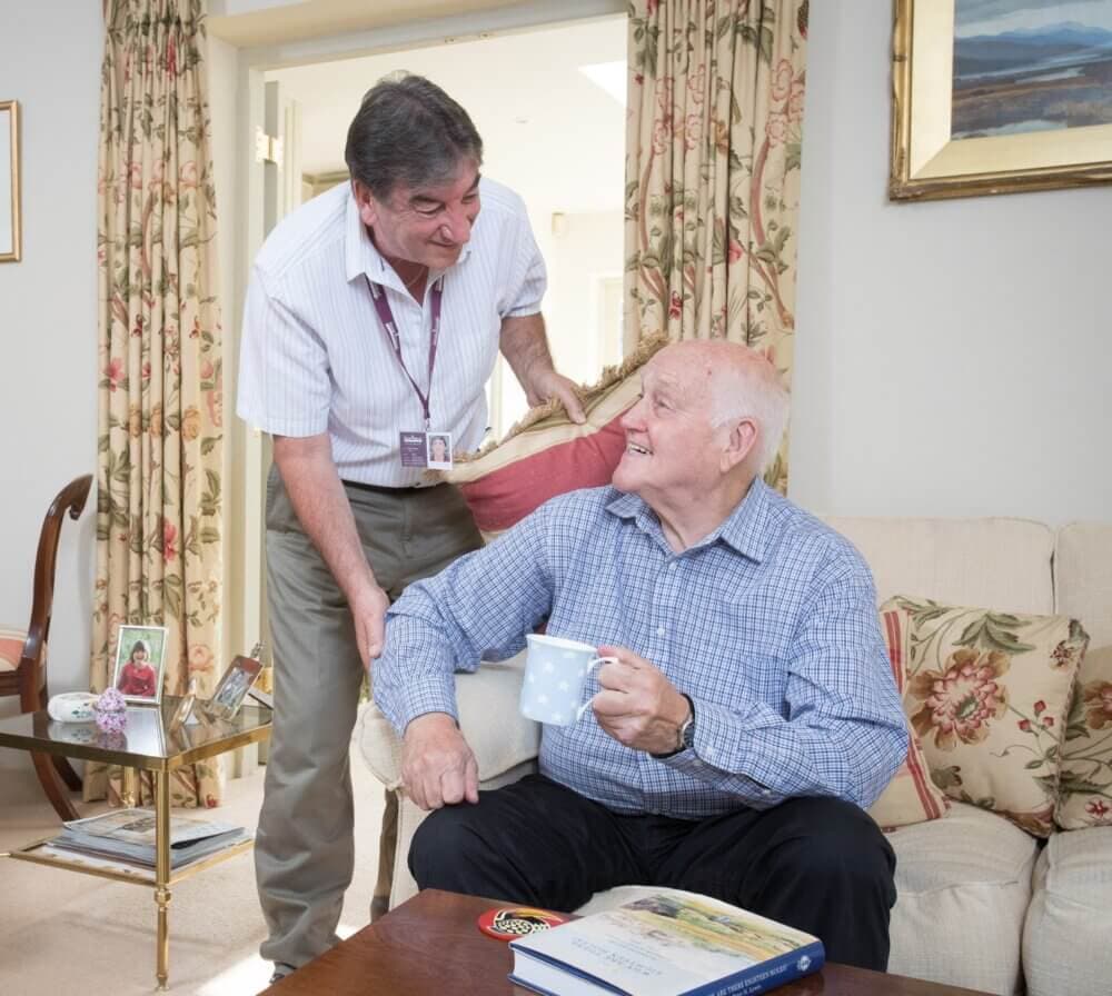 An elderly man with a mug sits on a couch while a caregiver smiles at him in a warmly decorated living room. - Home Instead