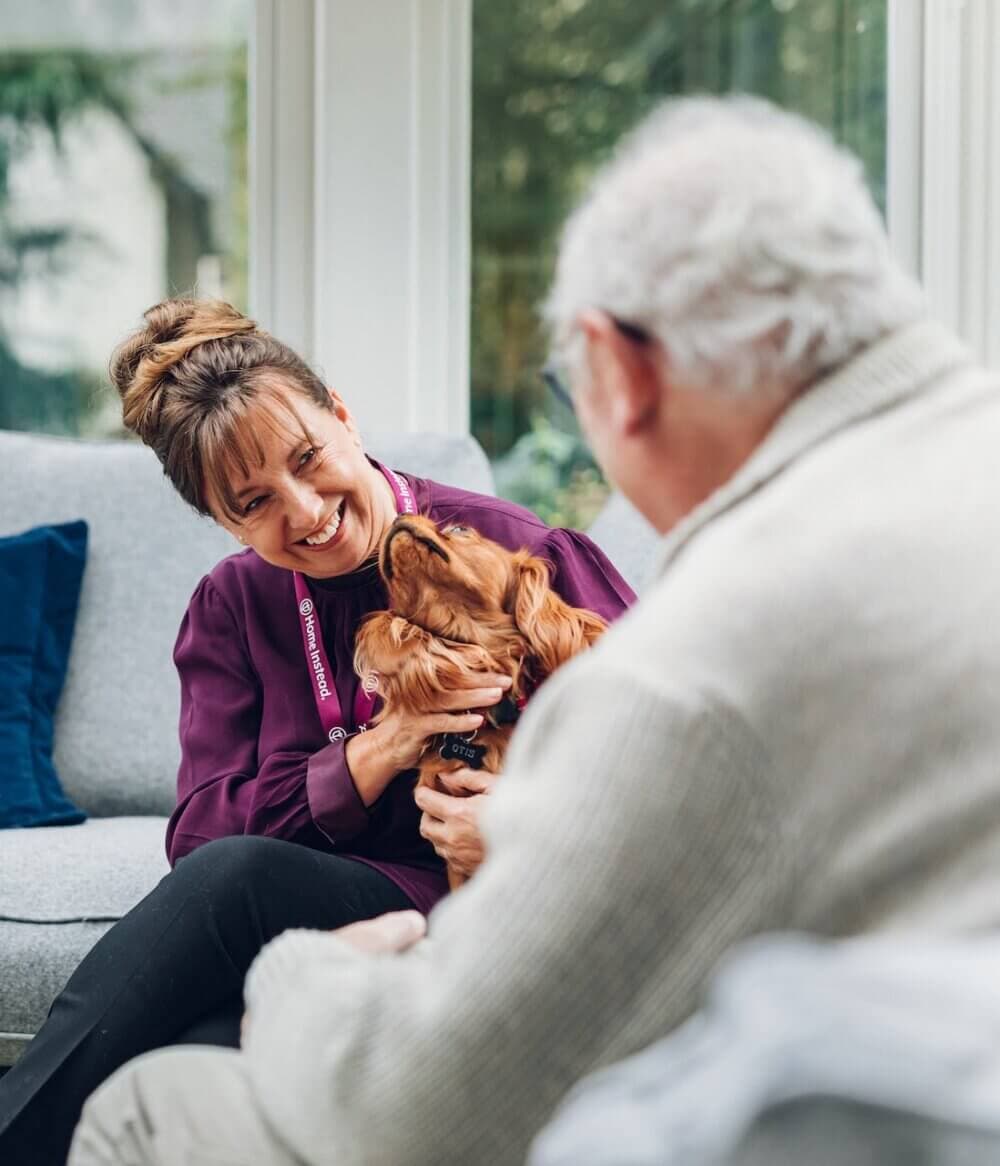A woman and elderly man smile at each other while the man holds a dog in a cosy living room setting. - Home Instead Southampton