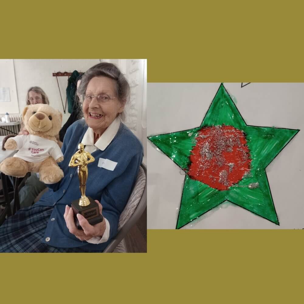 Elderly woman holding a teddy bear and a trophy next to a green star-shaped drawing with a red center. - Home Instead