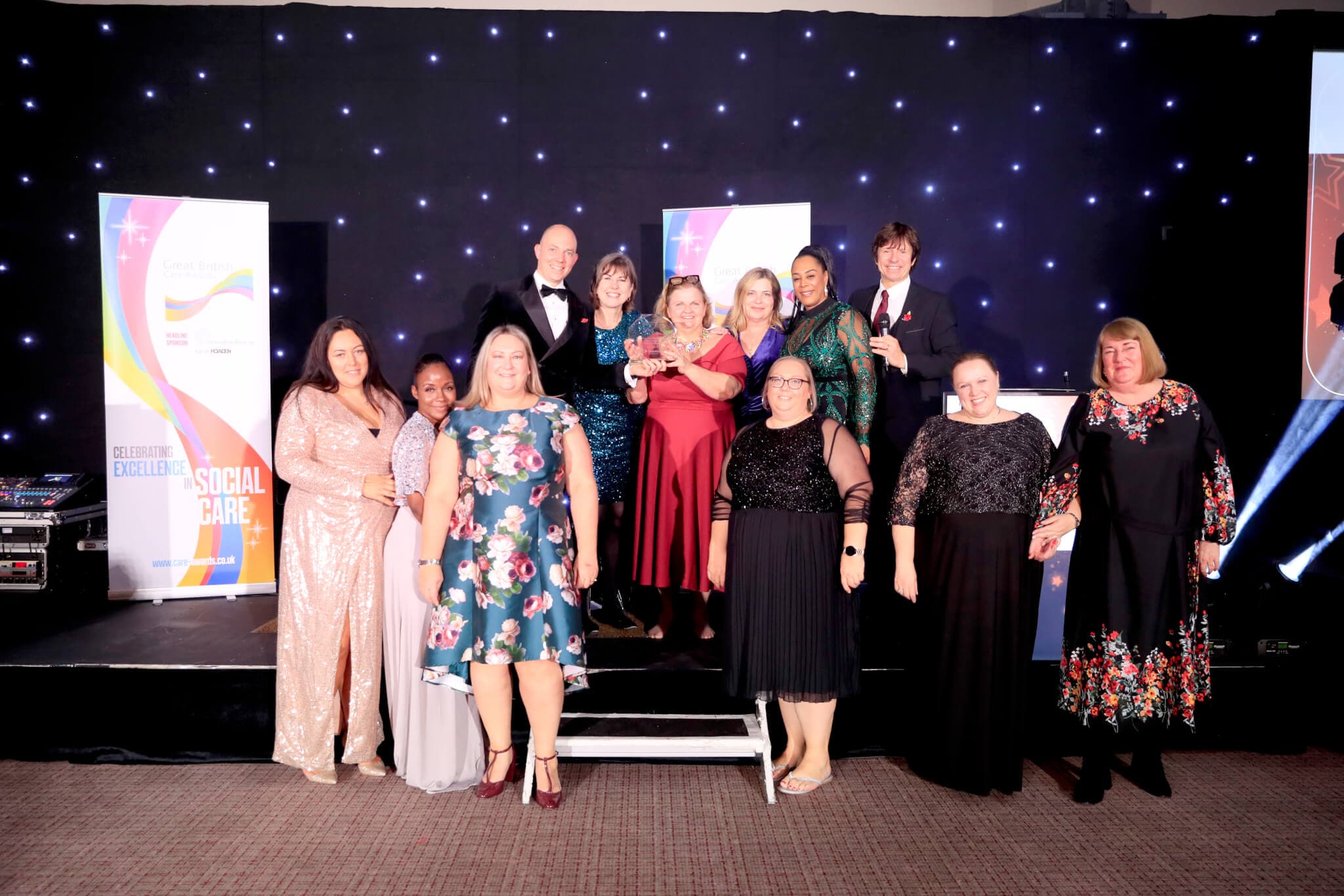 A group of people in formal attire posing for a photo on stage with twinkling star lights and award banners in the background. - Home Instead