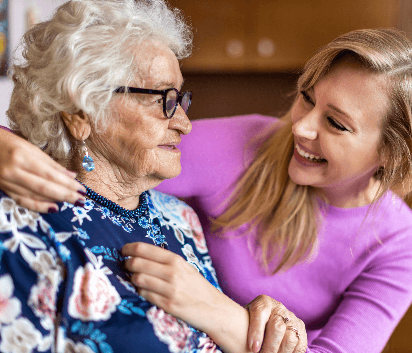 Elderly woman with grey hair and glasses embracing a smiling younger woman in a purple top. - Home Instead