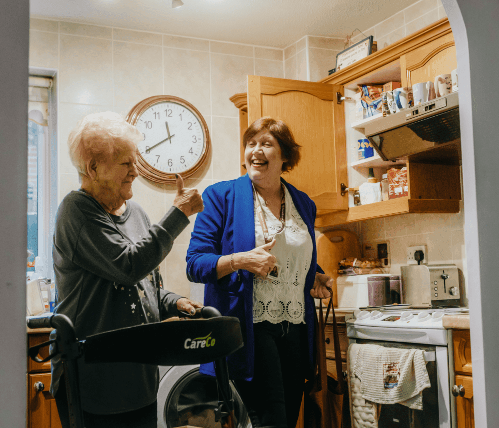 Two women laughing together in a kitchen, one elderly using a walking aid, the other holding a cup, with a large clock behind. - Home Instead