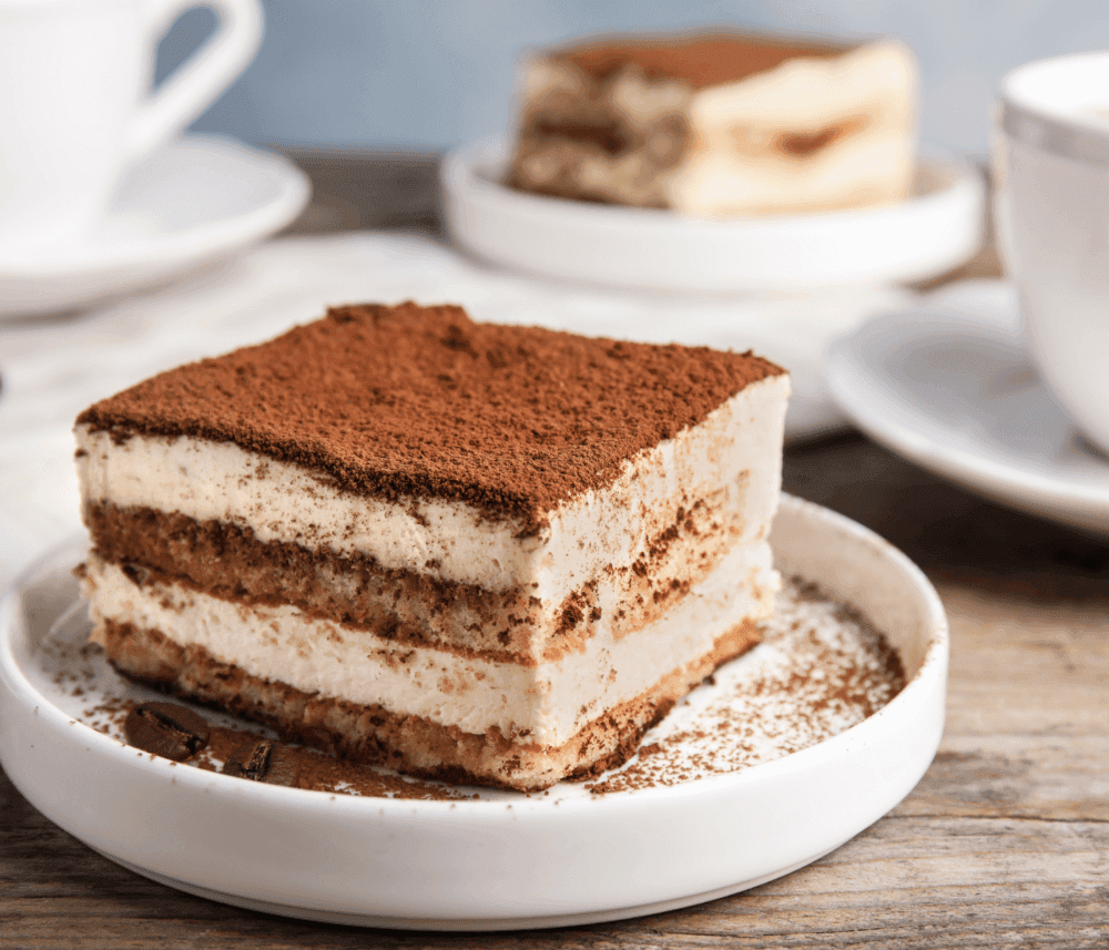 A slice of tiramisu on a white plate with a cup in the background on a wooden table. - Home Instead