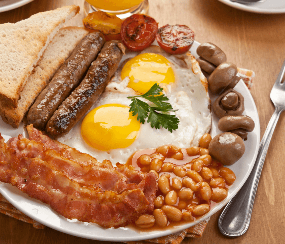 A plate with bacon, sausages, fried eggs, beans, tomatoes, mushrooms, and two slices of toast, set on a wooden table. - Home Instead