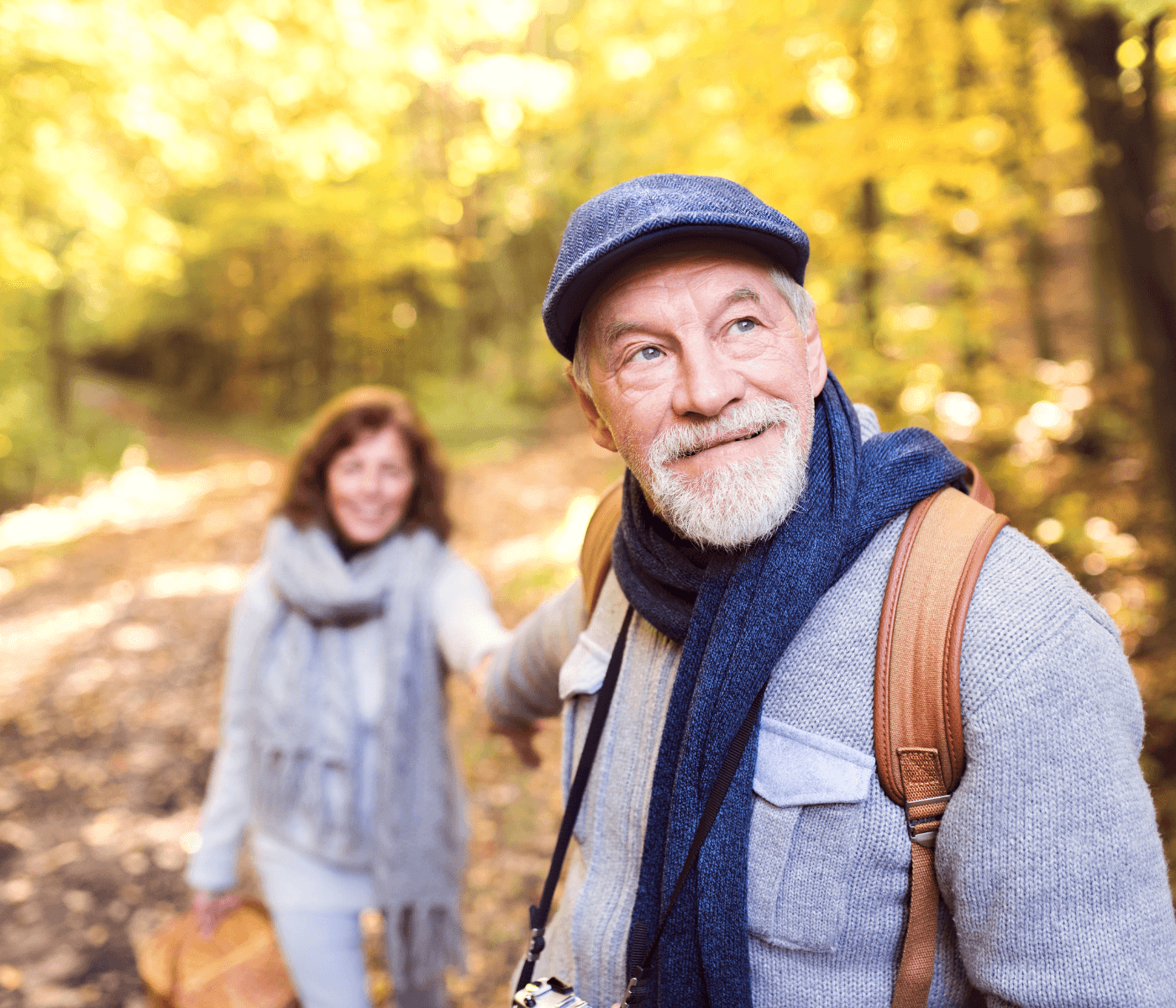 Elderly couple wearing scarves and walking in a sunlit autumn forest, with the man in focus and looking back. - Home Instead