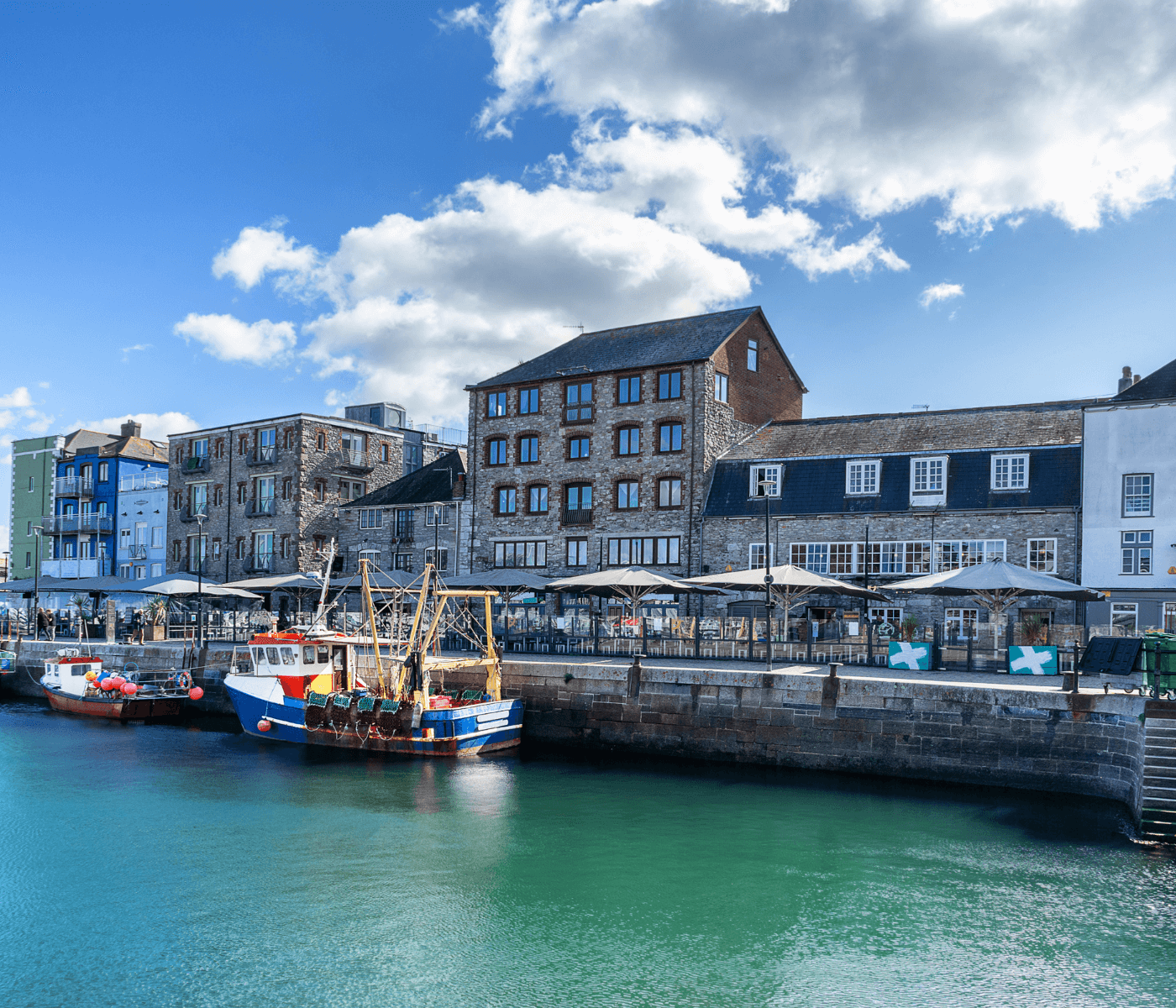 Colorful fishing boats docked in a vibrant, historic harbor with rustic buildings under a partly cloudy sky. - Home Instead