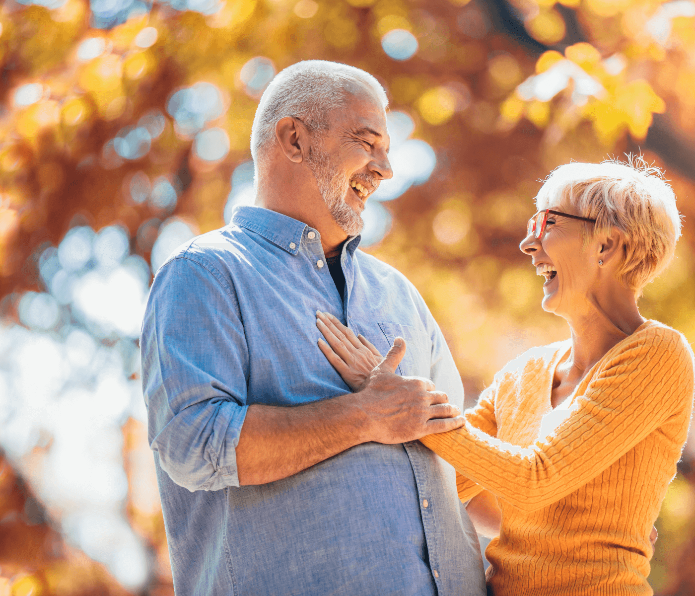 An older couple laughing and holding each other, standing outdoors with autumn foliage in the background. - Home Instead