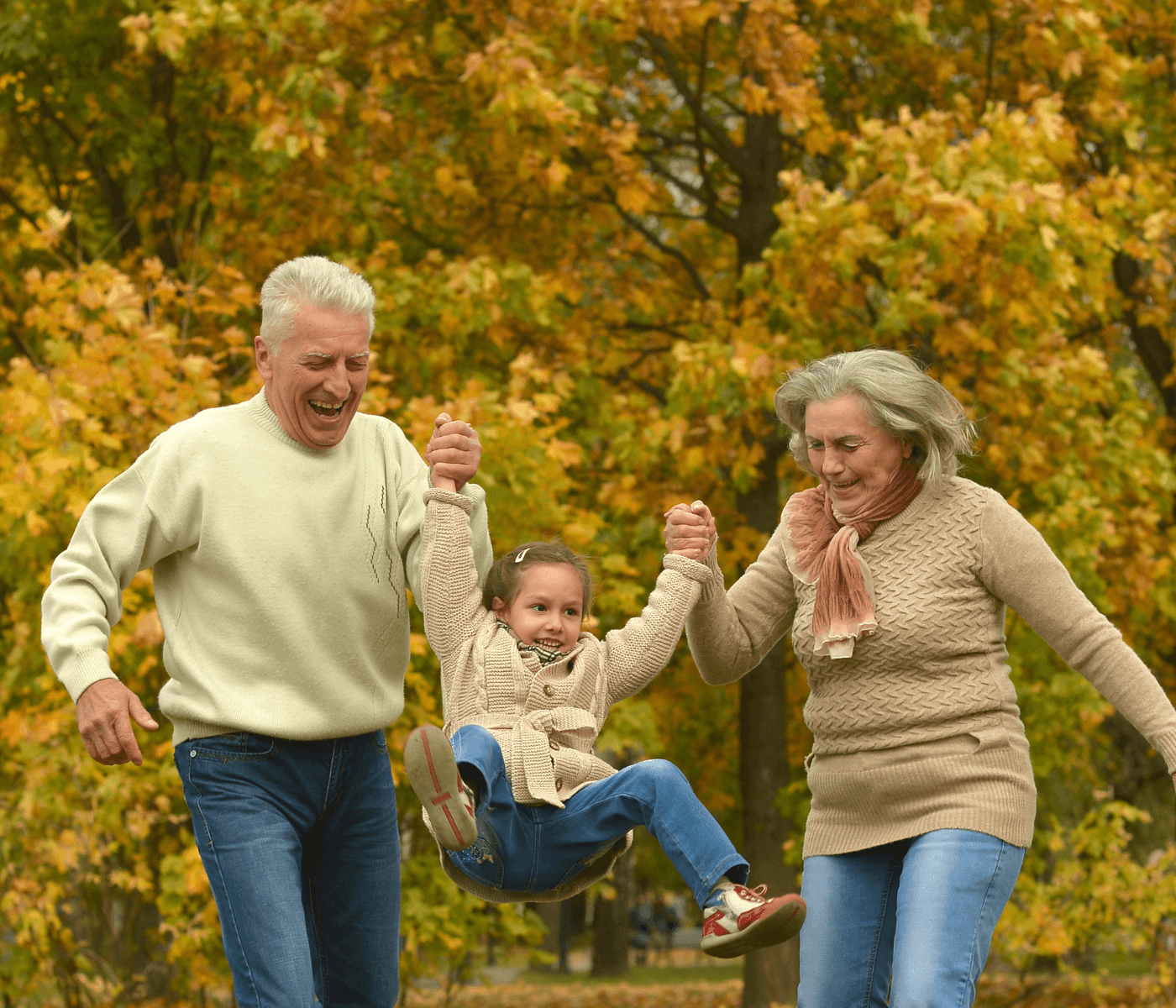 A happy child is being swung by two older adults in an autumn park with colorful leaves in the background. - Home Instead