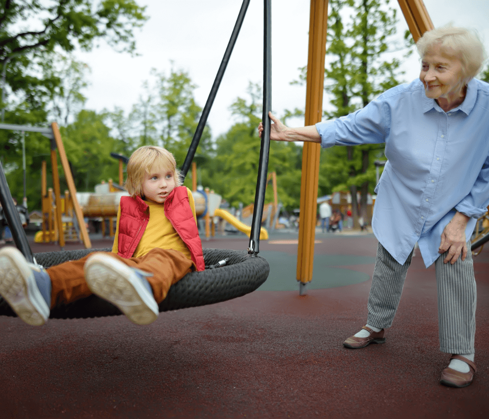 An elderly woman pushes a child on a swing at a playground, with trees and playground equipment in the background. - Home Instead
