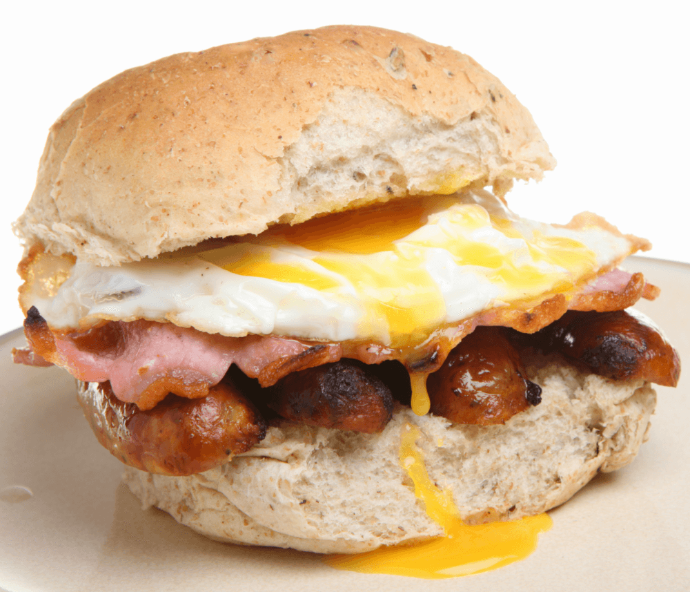 A sandwich with a fried egg, bacon, and sausages on a whole wheat bun. - Home Instead