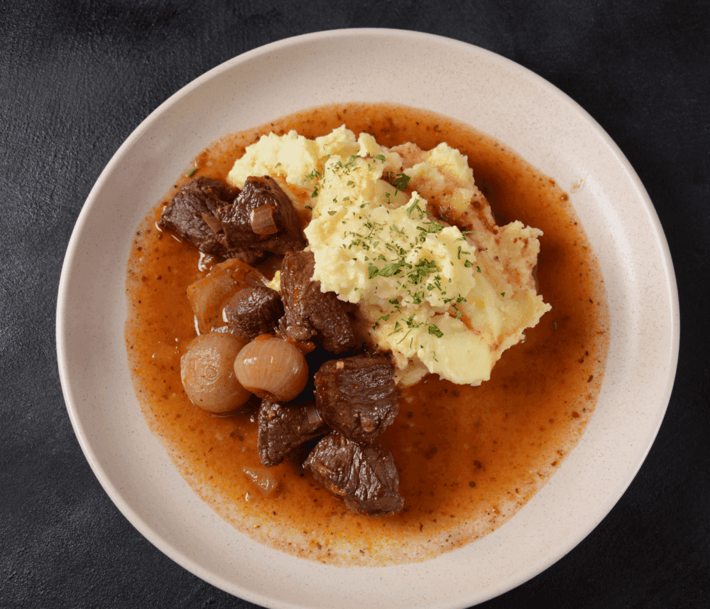 Plate of beef stew with onions, mashed potatoes topped with herbs, and rich brown sauce on a dark background. - Home Instead