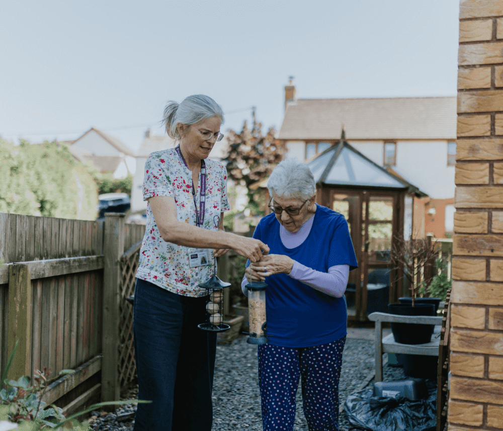 A caregiver assists an elderly woman in filling a bird feeder in a backyard garden on a sunny day. - Home Instead