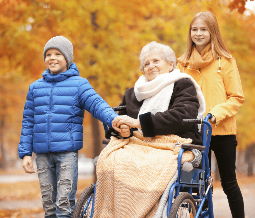 An elderly woman in a wheelchair is outside, with a boy holding her hand and a girl standing beside her. Autumn leaves surround them. - Home Instead