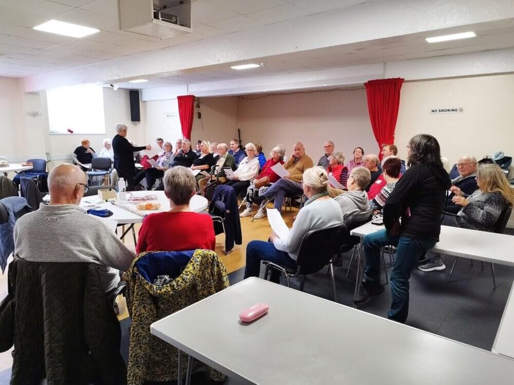 A group of older adults sit in a community hall, listening to a speaker in front of them. - Home Instead