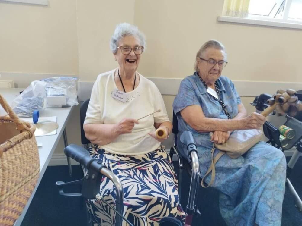 Two elderly women smiling and playing musical instruments while sitting on mobility scooters indoors. - Home Instead