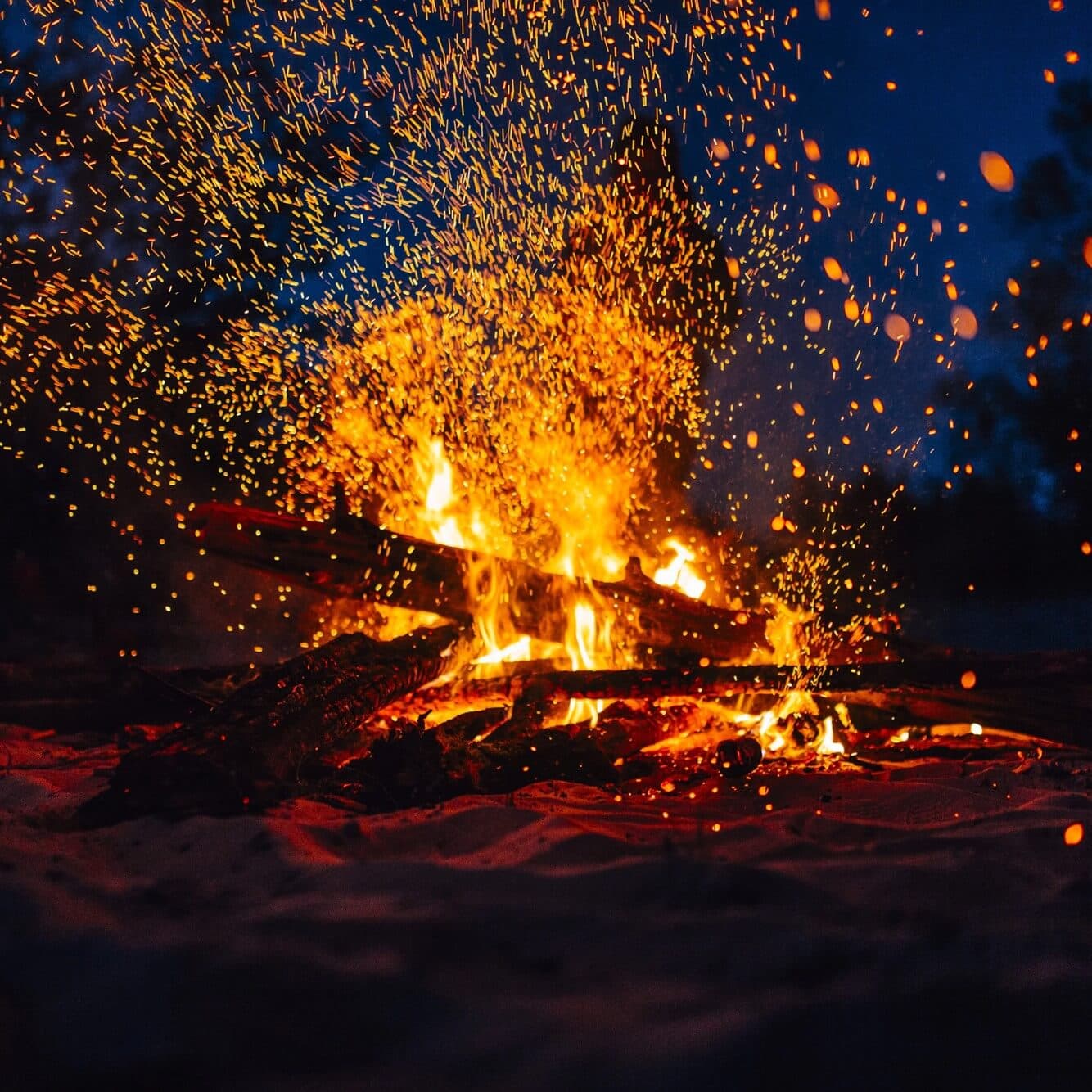 A campfire with glowing embers and sparks flying in the dark, set against a twilight sky with trees in the background. - Home Instead
