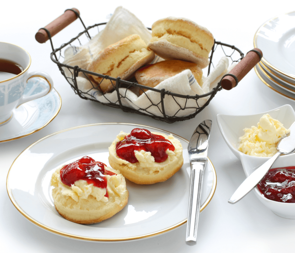 Tea with scones, clotted cream, and strawberry jam on a white table setting with a wire basket of scones in the background. - Home Instead