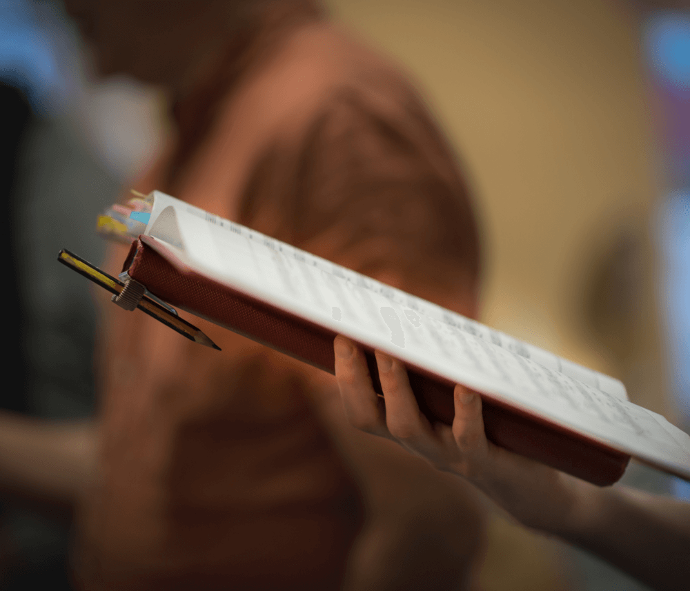 A person holds an open red binder with papers and a pencil attached, with a blurred background. - Home Instead