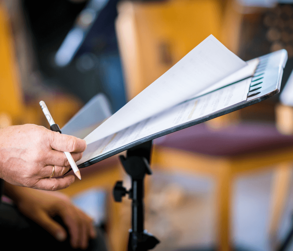 Close-up of a person's hand holding a pencil and flipping through sheet music on a music stand. - Home Instead